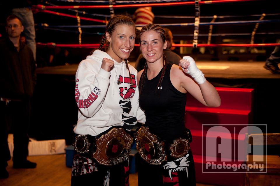 Two kickboxing women posing with fists clinche in front of a ring