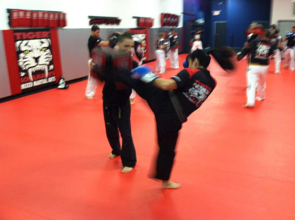 Woman exercising Kicking with coach with other fighters in the back at Tiger Schulmann's gym