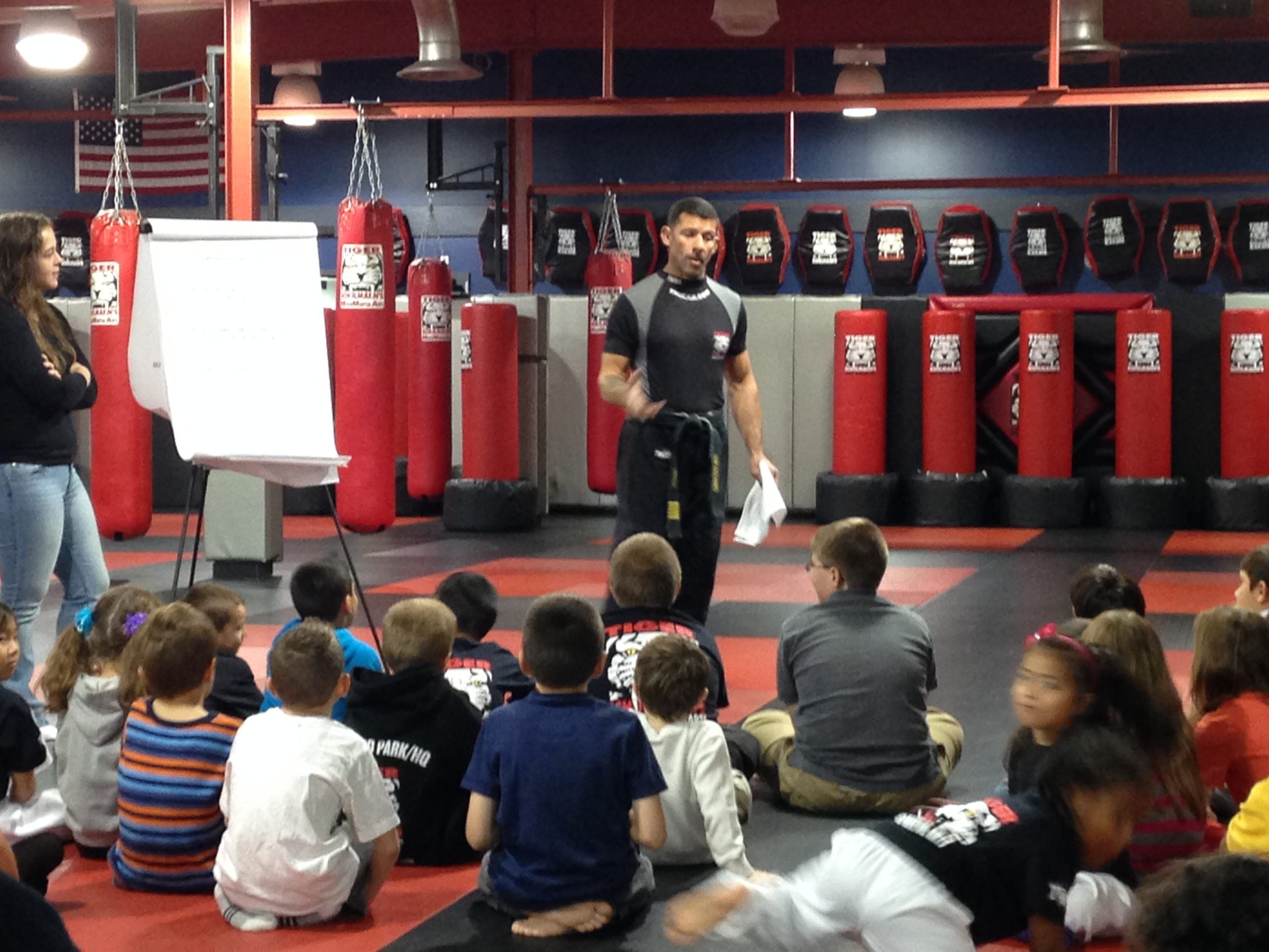 Kids sitting on the gym floor with their instructor standing and talking at Tiger Schulmann's