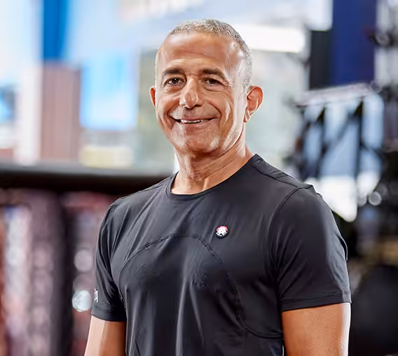 Tiger Schulmann smiles in a black shirt, standing confidently in a gym with blurred background elements.