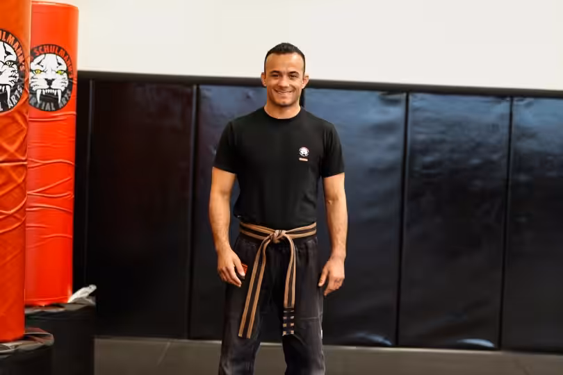 Man in black shirt and brown-striped belt stands smiling on black mat near red padded pillar with tiger logo