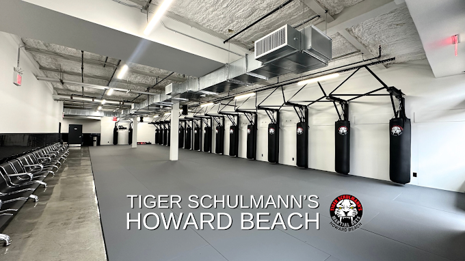 Martial arts gym with gray mats, black punching bags on frames, and rows of chairs. Text: TIGER SCHULMANN'S HOWARD BEACH & logo.