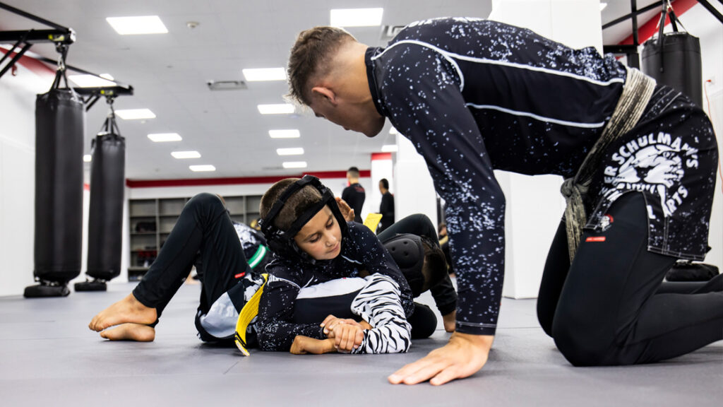 Two barefoot young fighters in black and white attire grapple on a gray mat. Referee watches from the right.