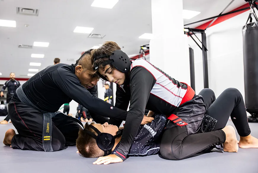 Instructor overseeing student applying submission hold to another student during grappling training in gym.