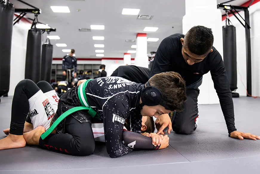 Instructor in black gi applying armlock submission to student wearing black headgear and green belt on gym mat.