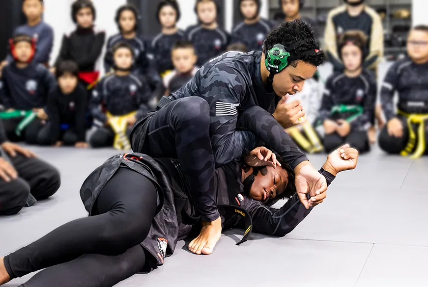 Instructor in black gi with green headgear demonstrating grappling technique on student while class watches on mat.